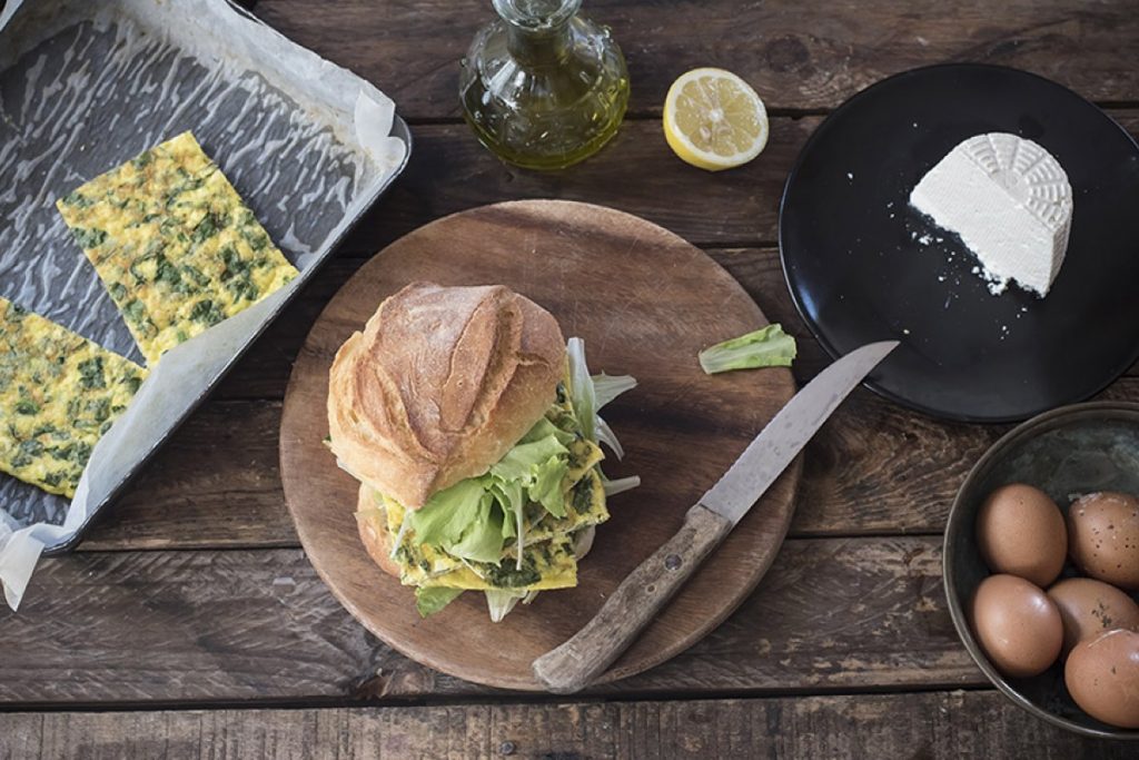 piatto scuro con un panino farcito di frittata e insalata, un coltello, uova e tranci di frittata