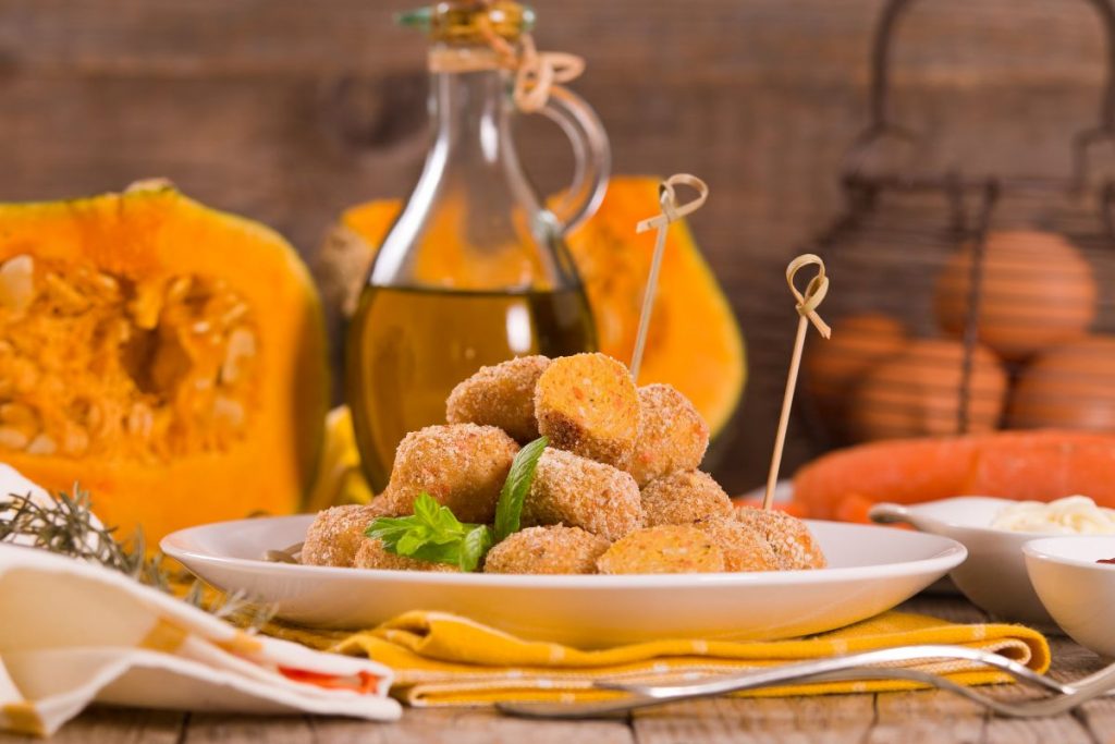 piatto bianco con polpette di zucca e spiedini, zucca aperta e flacone di olio sullo sfondo