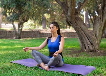 una ragazza seduta in posizione yoga sul tappetino su un prato, sotto un albero