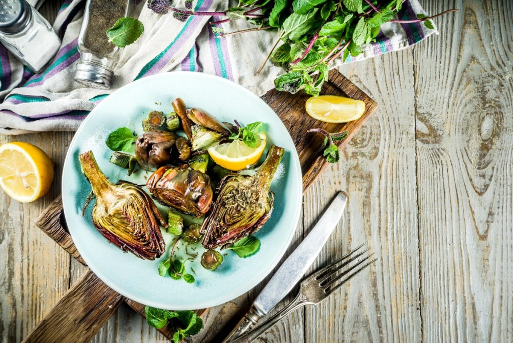 su tavolo di legno, piatto azzurro con 3 carciofi alla romana, forchetta e coltello accanto, mezzo lime e dietro sale e erbe aromatiche