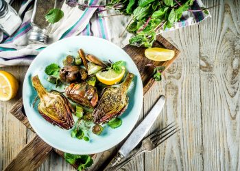 su tavolo di legno, piatto azzurro con 3 carciofi alla romana, forchetta e coltello accanto, mezzo lime e dietro sale e erbe aromatiche