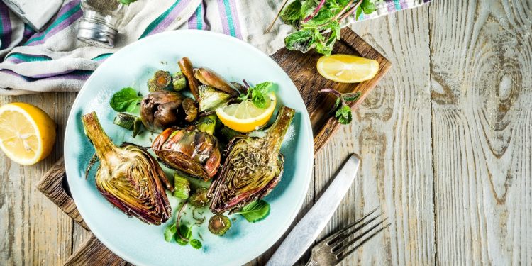 su tavolo di legno, piatto azzurro con 3 carciofi alla romana, forchetta e coltello accanto, mezzo lime e dietro sale e erbe aromatiche