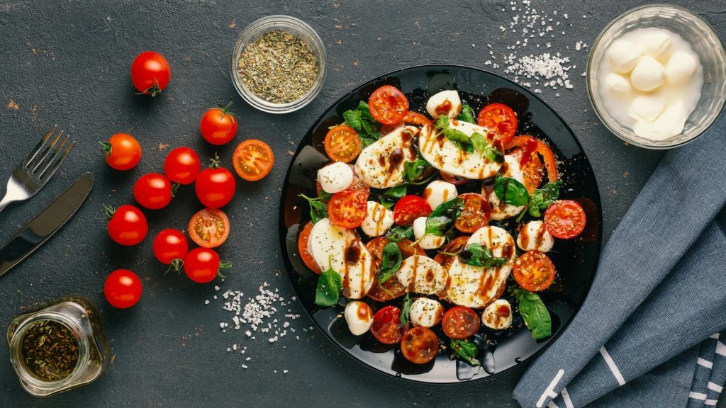 tavolo nero visto dall'alto con un piatto di insalata caprese e con ingredienti, sale, mozzarella, pomodori e origano