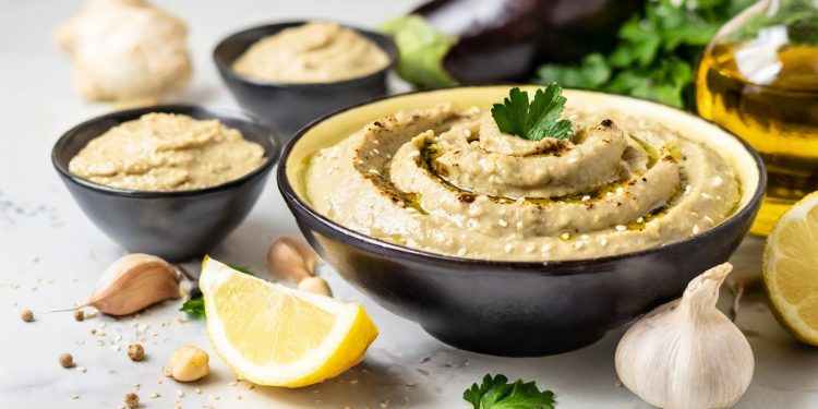 Piano da cucina, primo piano di una ciotola nera piena di babaganoush, attorno gli ingredienti della ricetta, aglio, limone, prezzemolo e melanzane