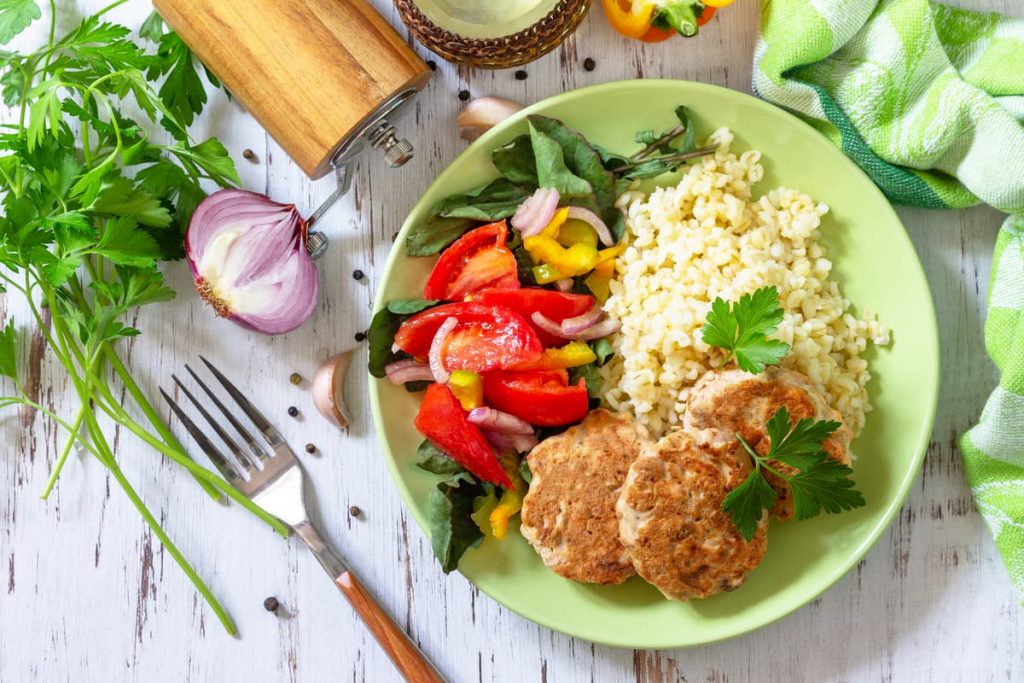 su tavolo bianco di legno un piatto verde con polpette di tonno, riso e peperoni. Attorno aglio, 1 forchetta e pepe macinato