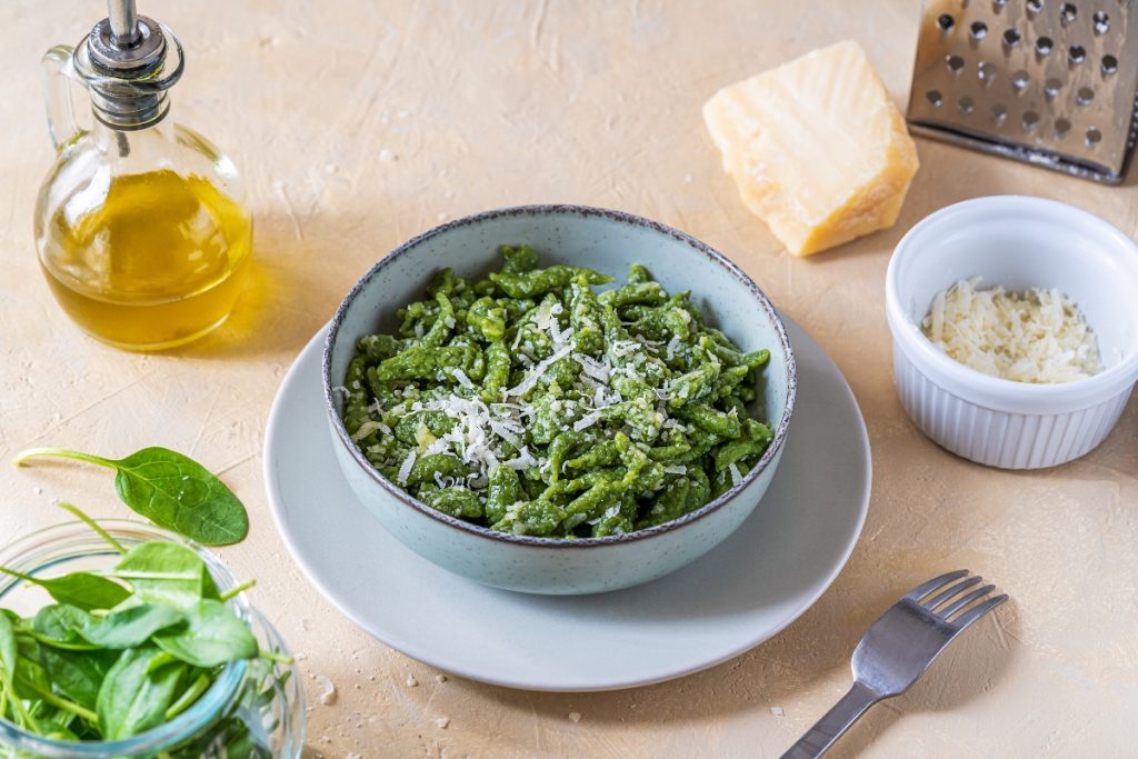 Su tavolo di legno chiaro ciotola con gli Spaetzle verdi fatti con gli spinaci, accanto spinaci freschi, una forchetta, olio EVO, parmigiano grattugiato