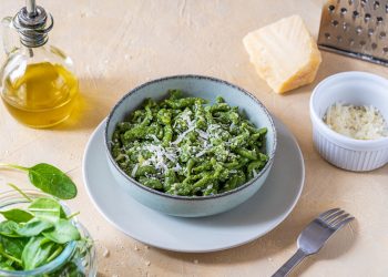 Su tavolo di legno chiaro ciotola con gli Spaetzle verdi fatti con gli spinaci, accanto spinaci freschi, una forchetta, olio EVO, parmigiano grattugiato