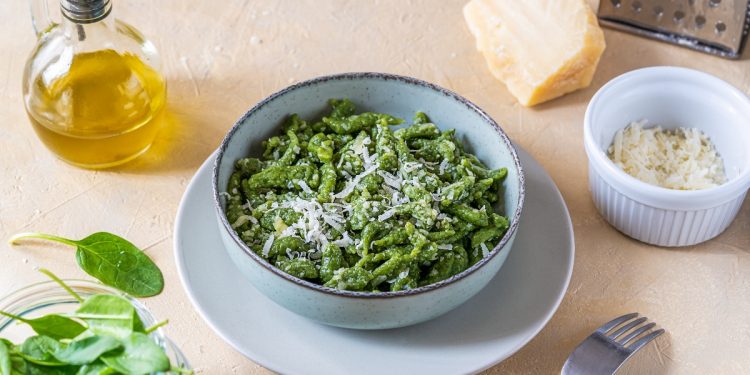 Su tavolo di legno chiaro ciotola con gli Spaetzle verdi fatti con gli spinaci, accanto spinaci freschi, una forchetta, olio EVO, parmigiano grattugiato