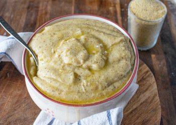 su tavola di legno, ciotola con la polenta taragna e un cucchiaio, accanto un bicchiere di farina di grano saraceno. Sotto la ciotola, c'è un canovaccio bianco