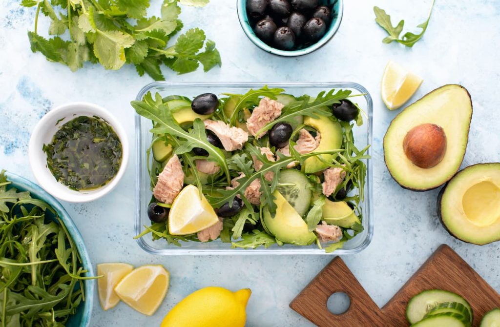 lunch box con insalata e tonno in scatola con attorno avocado, olive, limone