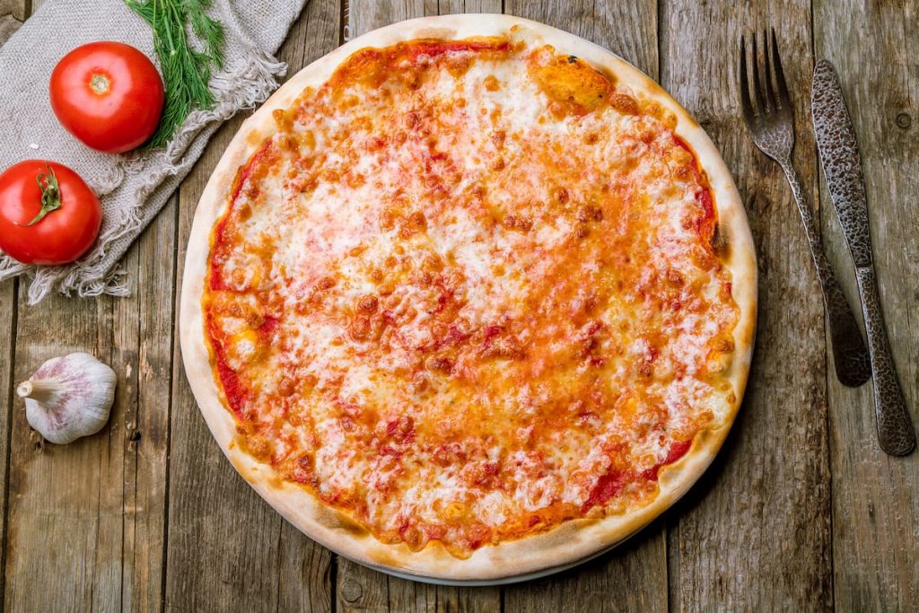 su un tavolo di legno una pizza margherita con forchetta e coltello, aglio e pomodori per far capire le calorie della pizza