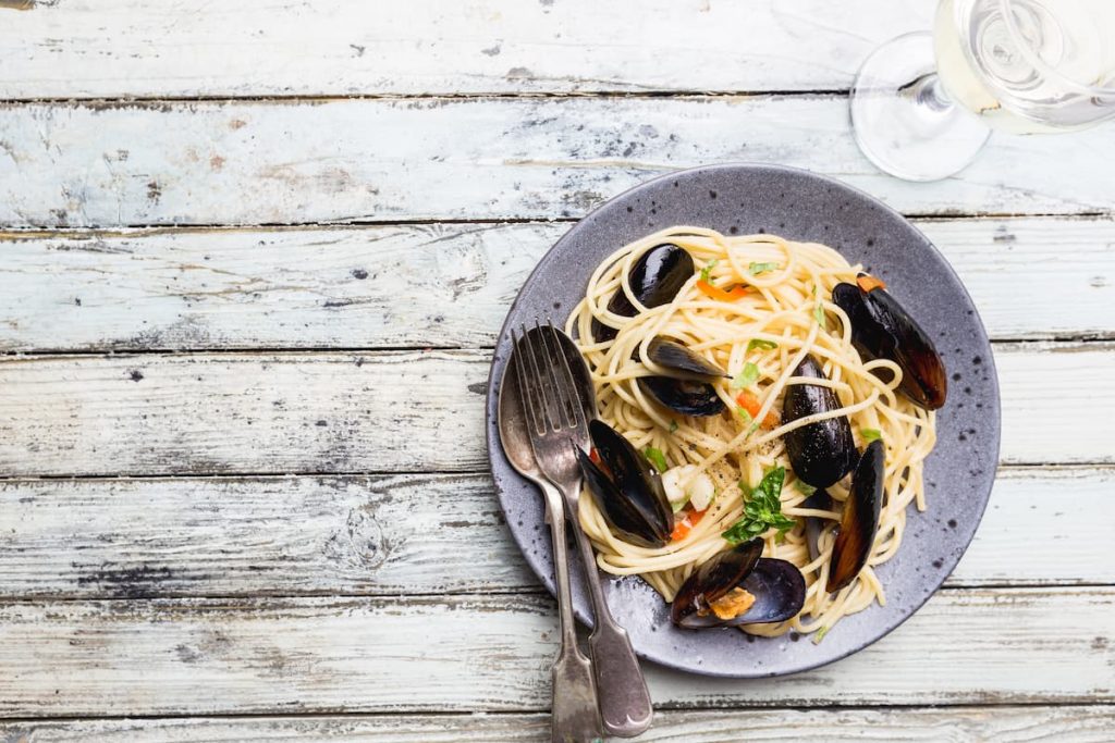 su un tavolo di legno bianco un piatto grigio con gli spaghetti con le cozze una forchetta e un cucchiaio