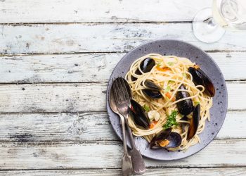 su un tavolo di legno bianco un piatto grigio con gli spaghetti con le cozze una forchetta e un cucchiaio