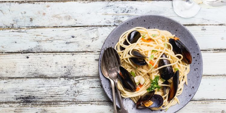 su un tavolo di legno bianco un piatto grigio con gli spaghetti con le cozze una forchetta e un cucchiaio