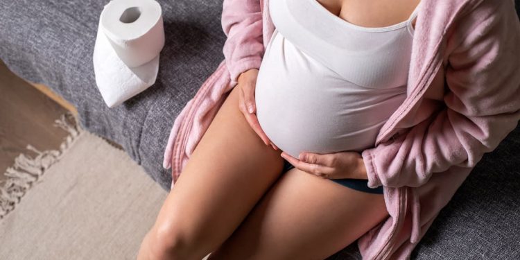 una donna in gravidanza seduta in bagno che si tiene la pancia perché soffre di stitichezza. Accanto a lei carta igienica