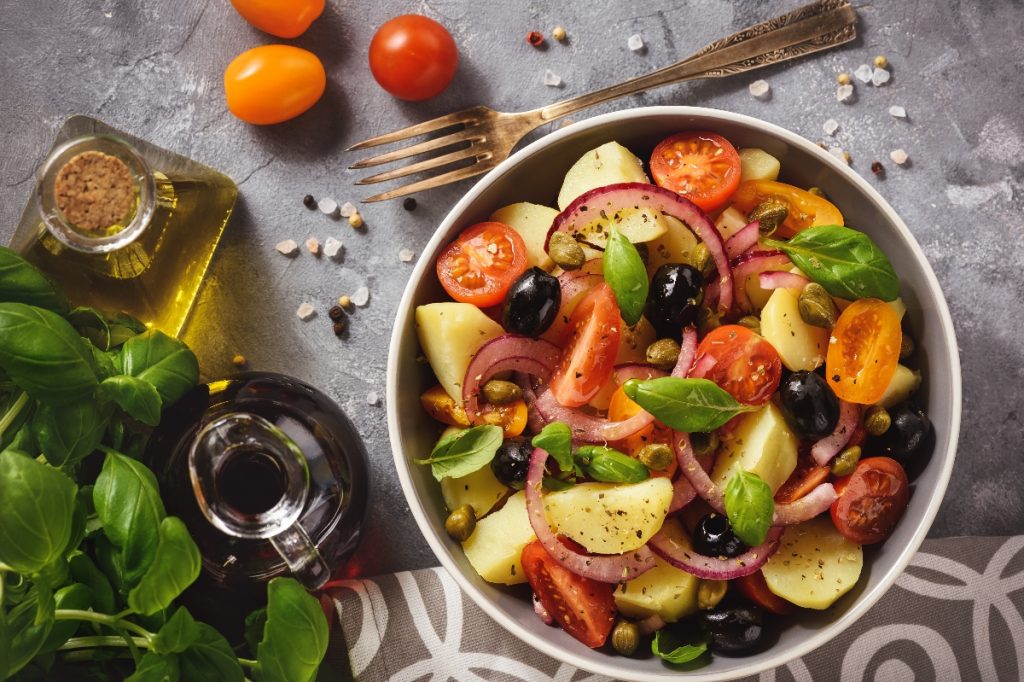 su tavolo una ciotola con l'insalata pantesca con patate pomodorini, capperi e cipolle rosse, accanto olio EVO e basilico