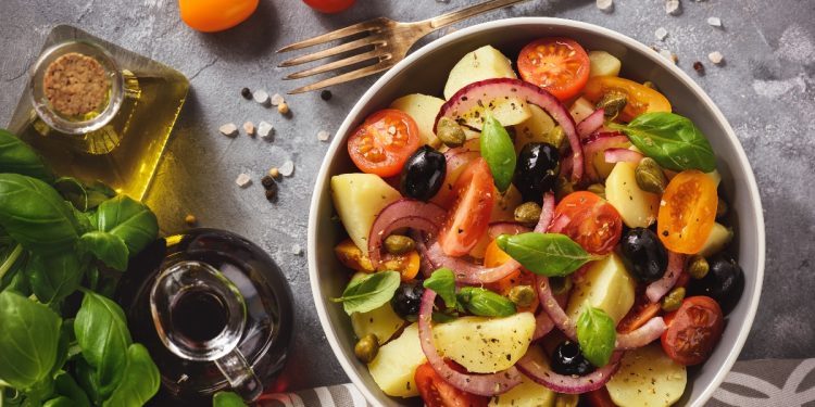su tavolo una ciotola con l'insalata pantesca con patate pomodorini, capperi e cipolle rosse, accanto olio EVO e basilico