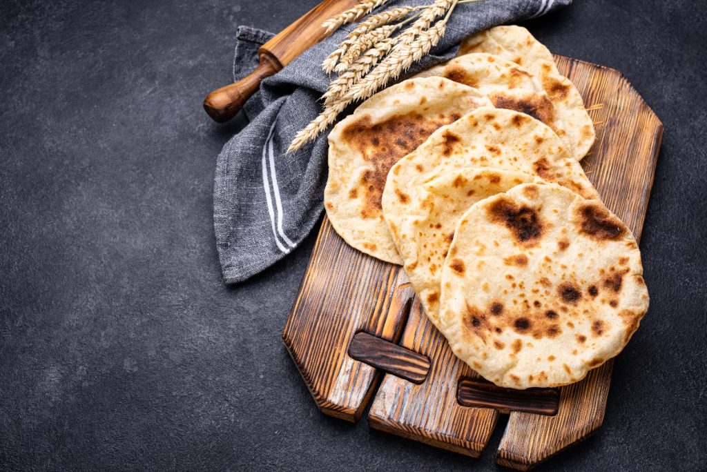 su sfondo nero, tagliere di legno con 4 pani pita, un mattarello