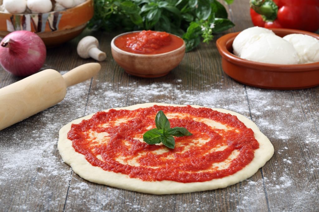 su piano da cucina di legno, pizza rossa cruda con passata di pomodori, un mattarello, cipolla, basilico, una ciotola con 2 mozzarelle