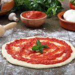 su piano da cucina di legno, pizza rossa cruda con passata di pomodori, un mattarello, cipolla, basilico, una ciotola con 2 mozzarelle