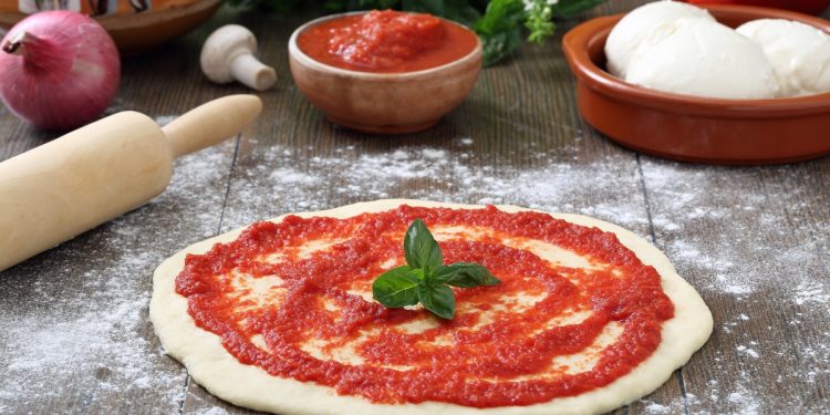 su piano da cucina di legno, pizza rossa cruda con passata di pomodori, un mattarello, cipolla, basilico, una ciotola con 2 mozzarelle