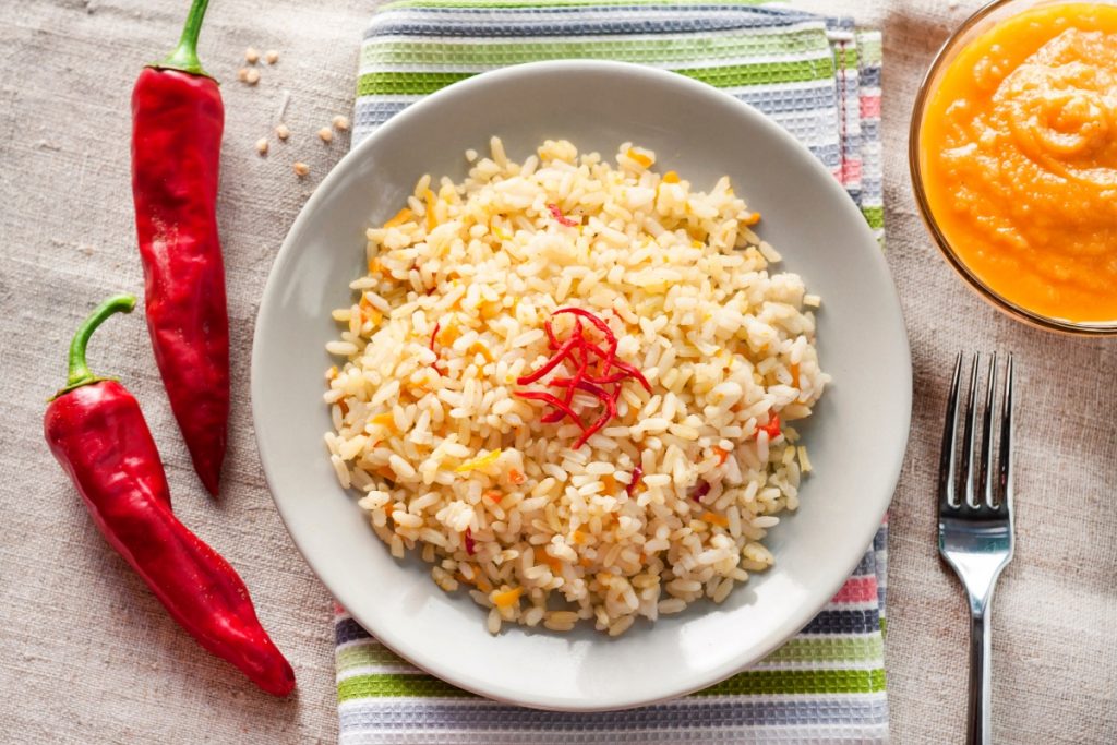 su tavolo con tovaglia beige piatto bianco con riso parboiled, 2 peperonicini, una ciotola con purée di carote e una forchetta