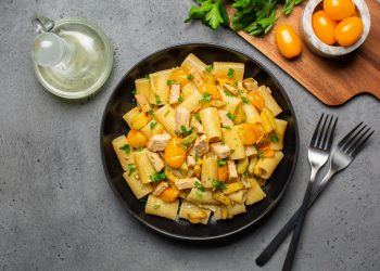 su tavolo grigio, piatto nero con pasta con pesce spada, prezzemolo e pomodorini gialli. Accanto una forchetta e un coltello e dietro un tagliere con pomodorini gialli e prezzemolo