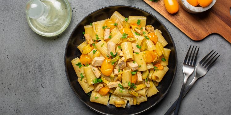 su tavolo grigio, piatto nero con pasta con pesce spada, prezzemolo e pomodorini gialli. Accanto una forchetta e un coltello e dietro un tagliere con pomodorini gialli e prezzemolo