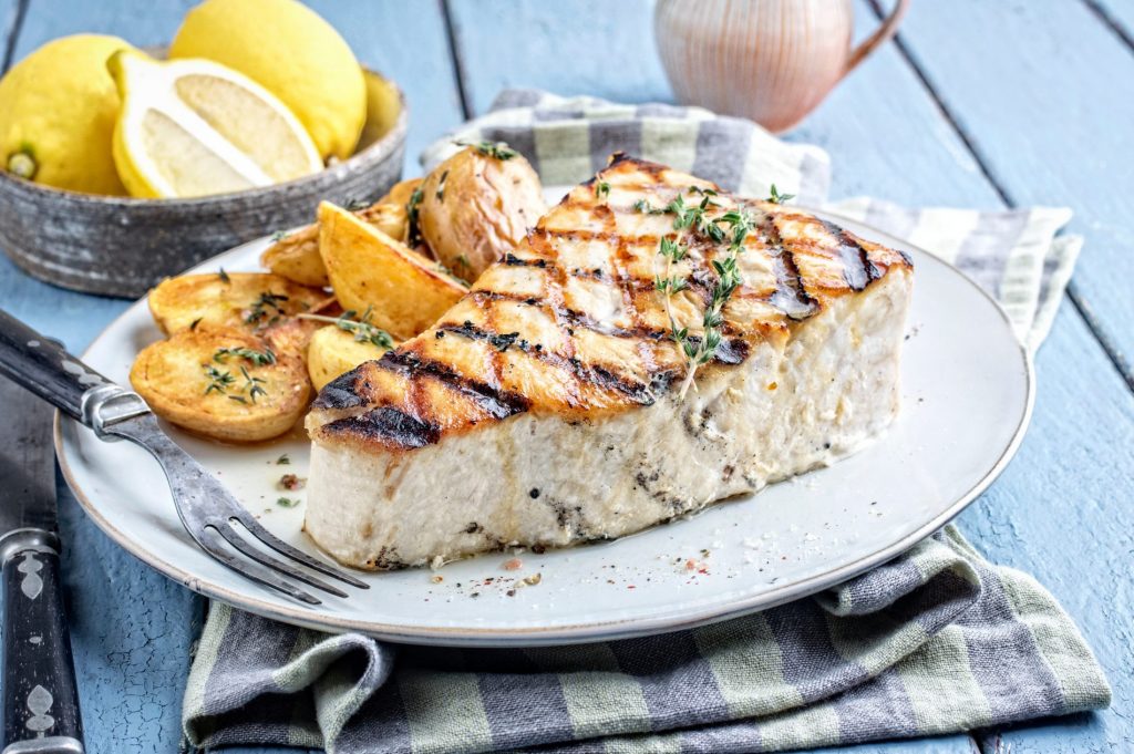 su un tavolo di legno blu, piatto bianco con pesce spada, un tovagliolo blu e bianco e fette di limoni