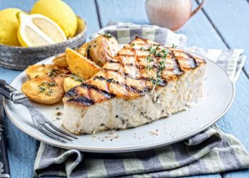 su un tavolo di legno blu, piatto bianco con pesce spada, un tovagliolo blu e bianco e fette di limoni