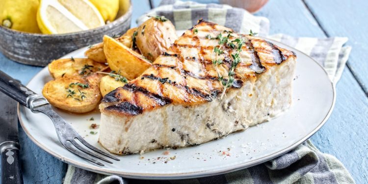 su un tavolo di legno blu, piatto bianco con pesce spada, un tovagliolo blu e bianco e fette di limoni