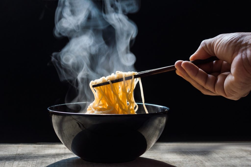 su tavolo ciotola nera con noodles fatti in casa che fumano, una mano li prende con le bacchette cinesi