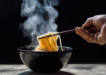 su tavolo ciotola nera con noodles fatti in casa che fumano, una mano li prende con le bacchette cinesi