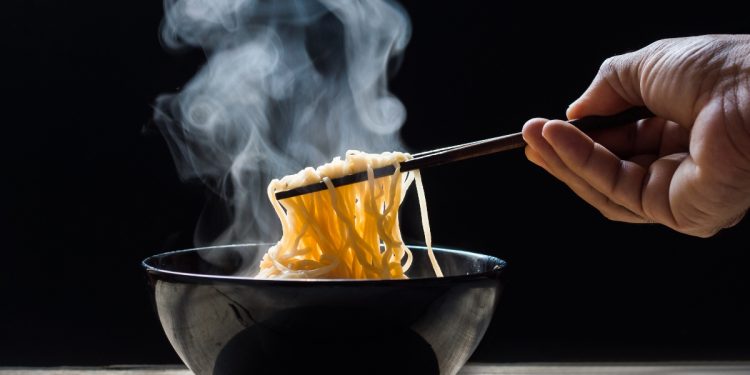 su tavolo ciotola nera con noodles fatti in casa che fumano, una mano li prende con le bacchette cinesi