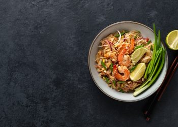 su sfondo nero un piatto bianco con un pad thai con gamberi, tofu e noodles. Accanto mezzo limone