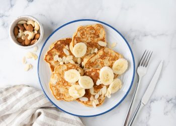 su piano di marmo, piatto con pancake proteici e banane tagliata, accanto forchetta e coltello e frutta secca