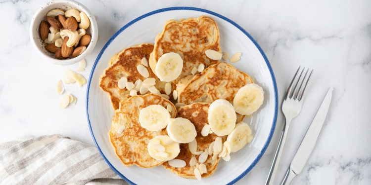 su piano di marmo, piatto con pancake proteici e banane tagliata, accanto forchetta e coltello e frutta secca