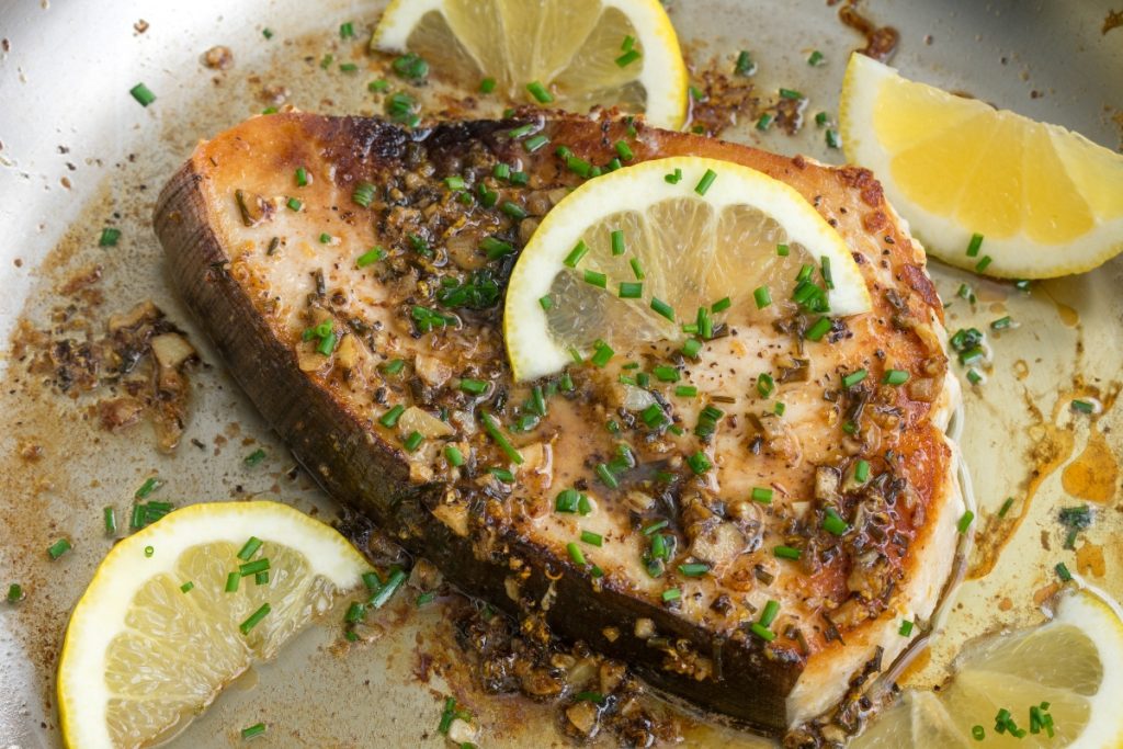 primo piano di una fetta di pesce spada in padella con erbe aromatiche e fette di limone