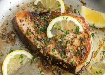 primo piano di una fetta di pesce spada in padella con erbe aromatiche e fette di limone