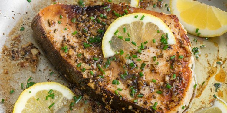 primo piano di una fetta di pesce spada in padella con erbe aromatiche e fette di limone