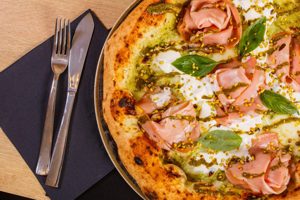 primo piano di una pizza mortadella e pistacchi con mozzarella su un tavolo e accanto tovagliolo di carta blu con forchetta e coltello