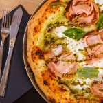 primo piano di una pizza mortadella e pistacchi con mozzarella su un tavolo e accanto tovagliolo di carta blu con forchetta e coltello