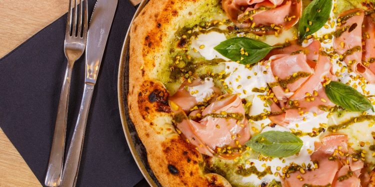 primo piano di una pizza mortadella e pistacchi con mozzarella su un tavolo e accanto tovagliolo di carta blu con forchetta e coltello