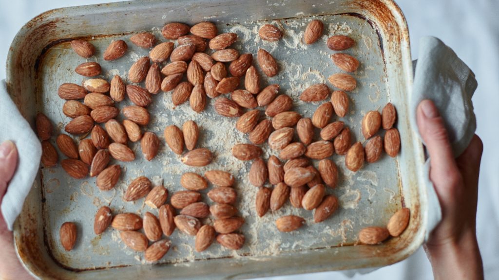 primo piano di mani che tengono una teglia da forno con carta da forno e mandorle tostate