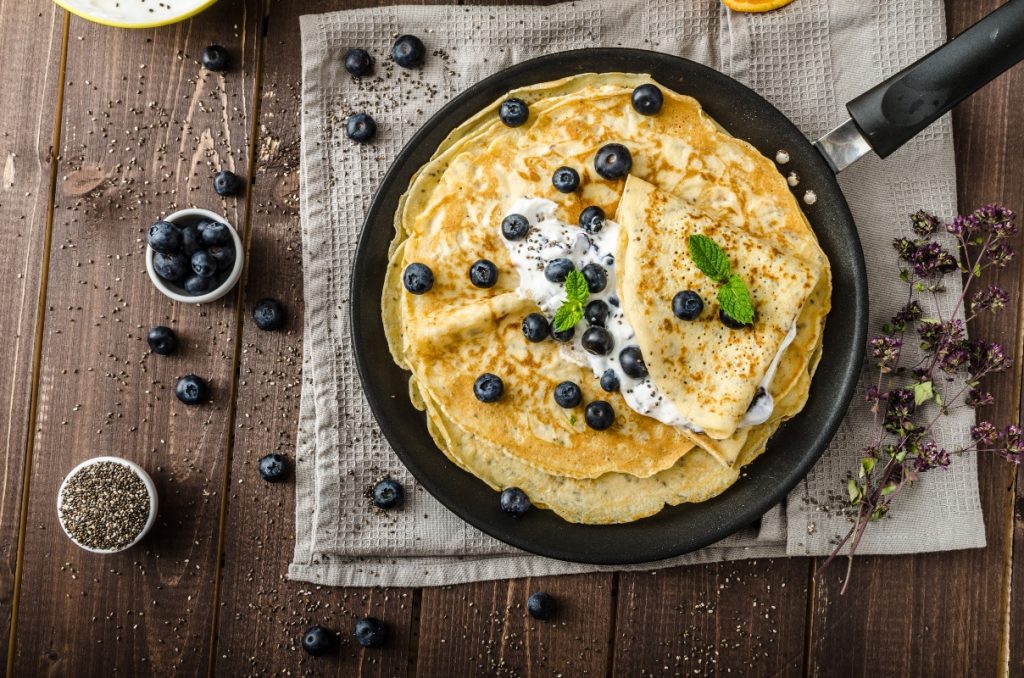 su tavolo di legno, canovaccio marrone con una padella e diverse crepes senza glutine con sopra dei mirtilli e accanto una ciotola piccola con mirtilli