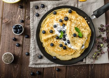 su tavolo di legno, canovaccio marrone con una padella e diverse crepes senza glutine con sopra dei mirtilli e accanto una ciotola piccola con mirtilli