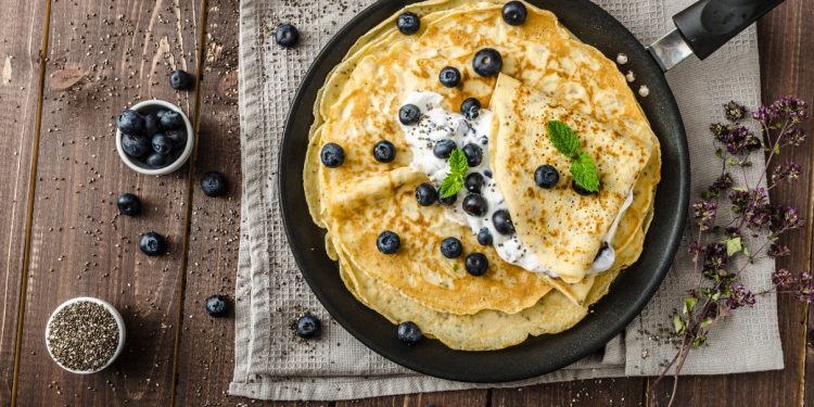 su tavolo di legno, canovaccio marrone con una padella e diverse crepes senza glutine con sopra dei mirtilli e accanto una ciotola piccola con mirtilli