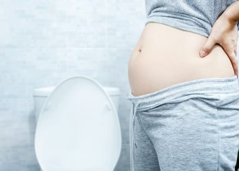 Primo piano di una donna con leggings e maglietta alzata con la pancia gonfia vicino la water perché soffre di intestino pigro