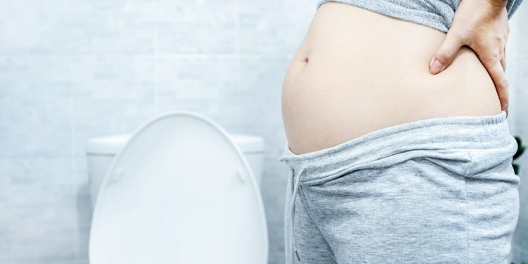 Primo piano di una donna con leggings e maglietta alzata con la pancia gonfia vicino la water perché soffre di intestino pigro