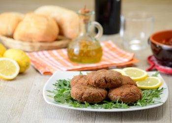 su tavolo da cucina, piatto con rucola, limoni tagliati e mondeghili, dietro una bottiglietta di olio EVO e panini